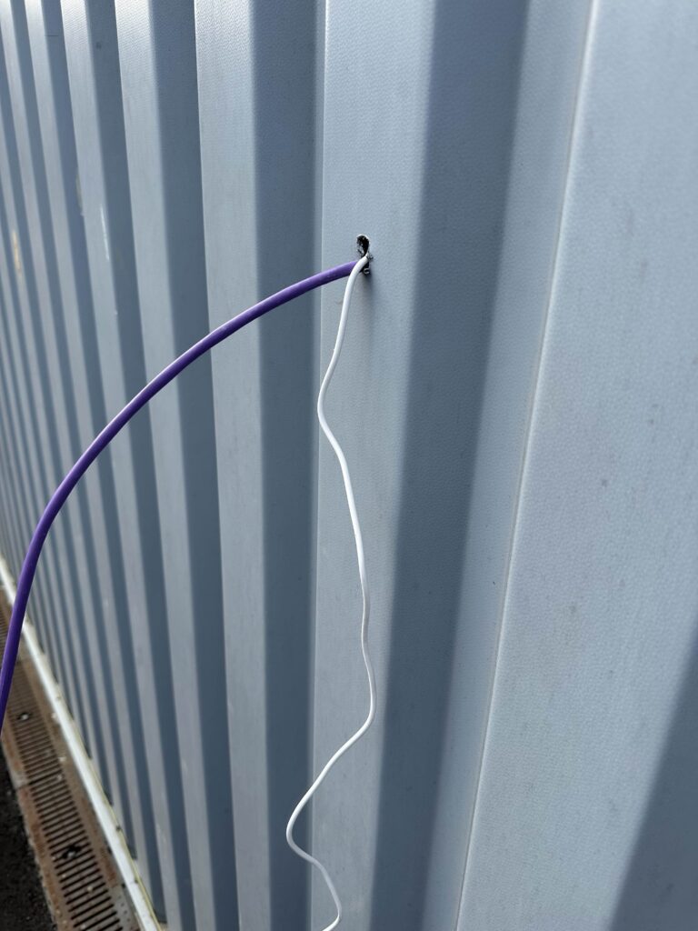 A purple ethernet network cable and a white USB-C power cable emerging from a drilled hole in a corrugated grey metal wall, the first step in solving a Faraday cage Wi-Fi issue.